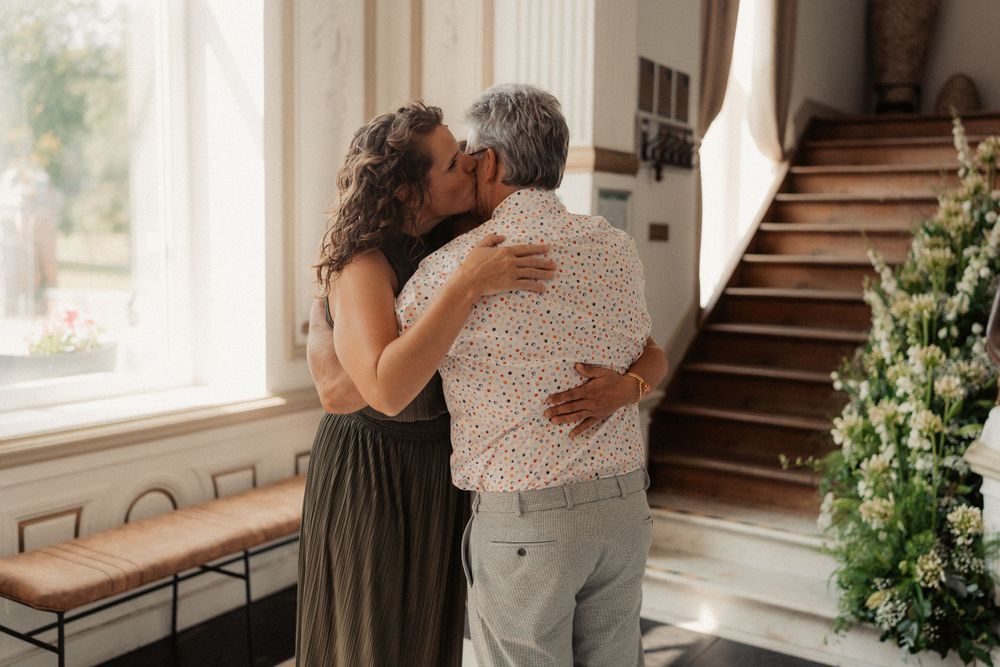 Couple sharing an intimate embrace by a window in a sunny interior setting with wooden staircase.