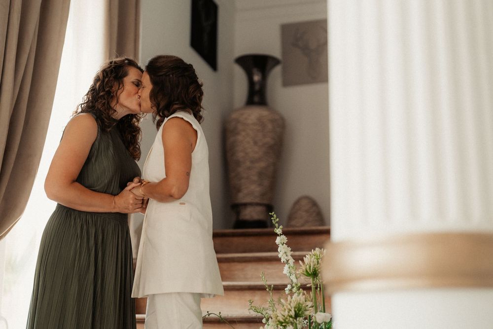 Two people share an intimate embrace in front of a decorative vase and white curtains.