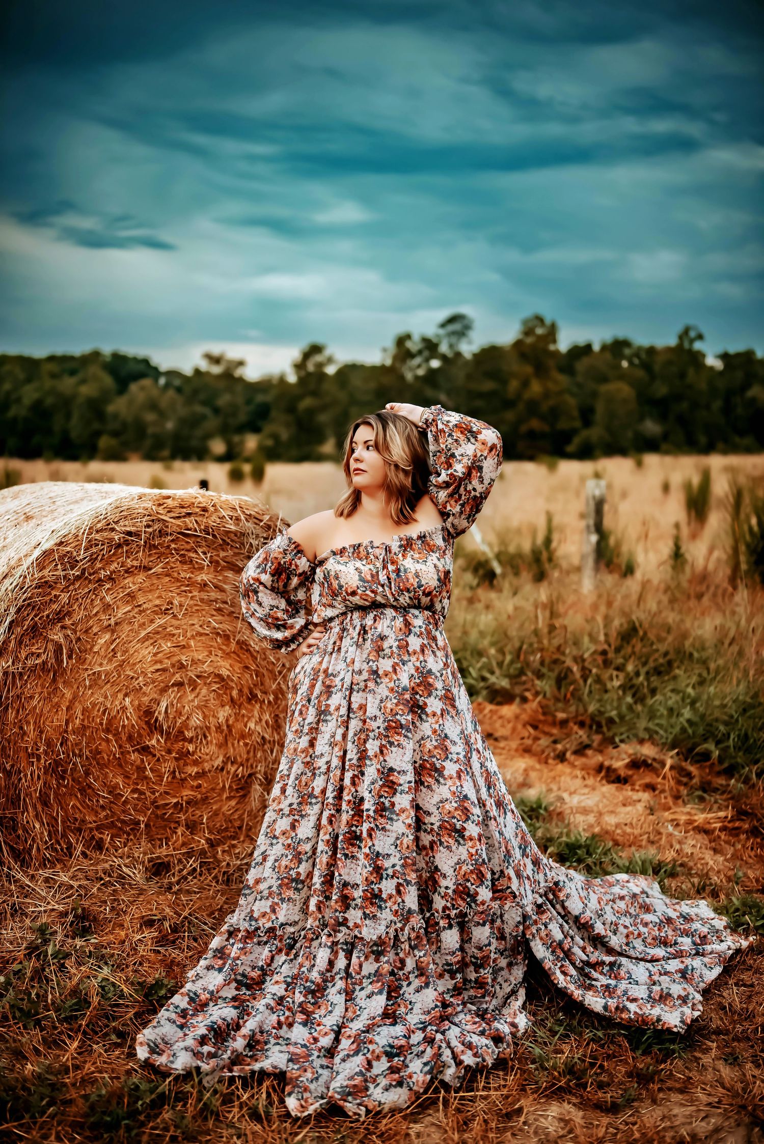 A floral maxi dress flows gracefully against a hay bale in a rustic field under moody teal skies.