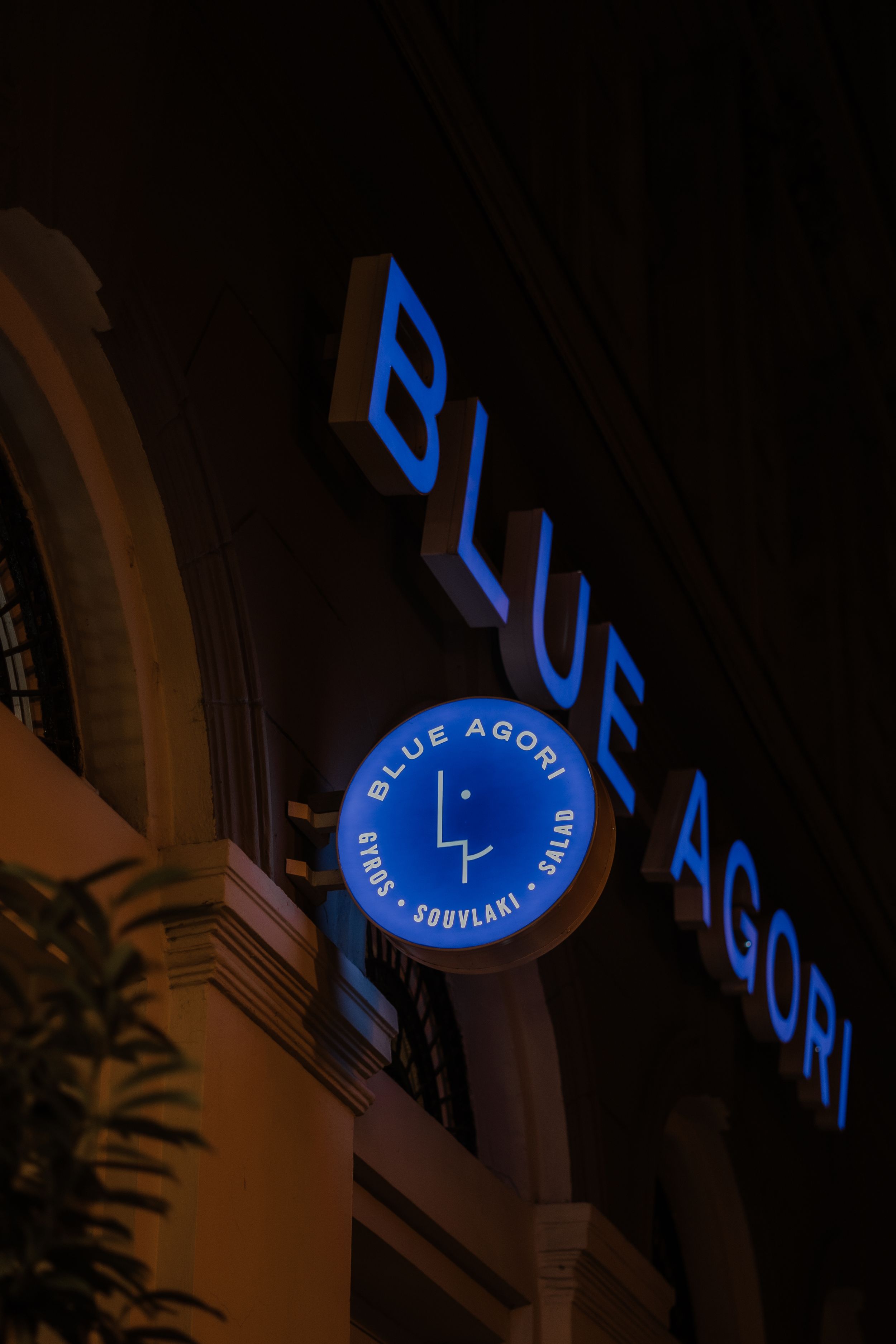 Illuminated blue sign and logo for Blue Agori displayed at night on a building exterior.