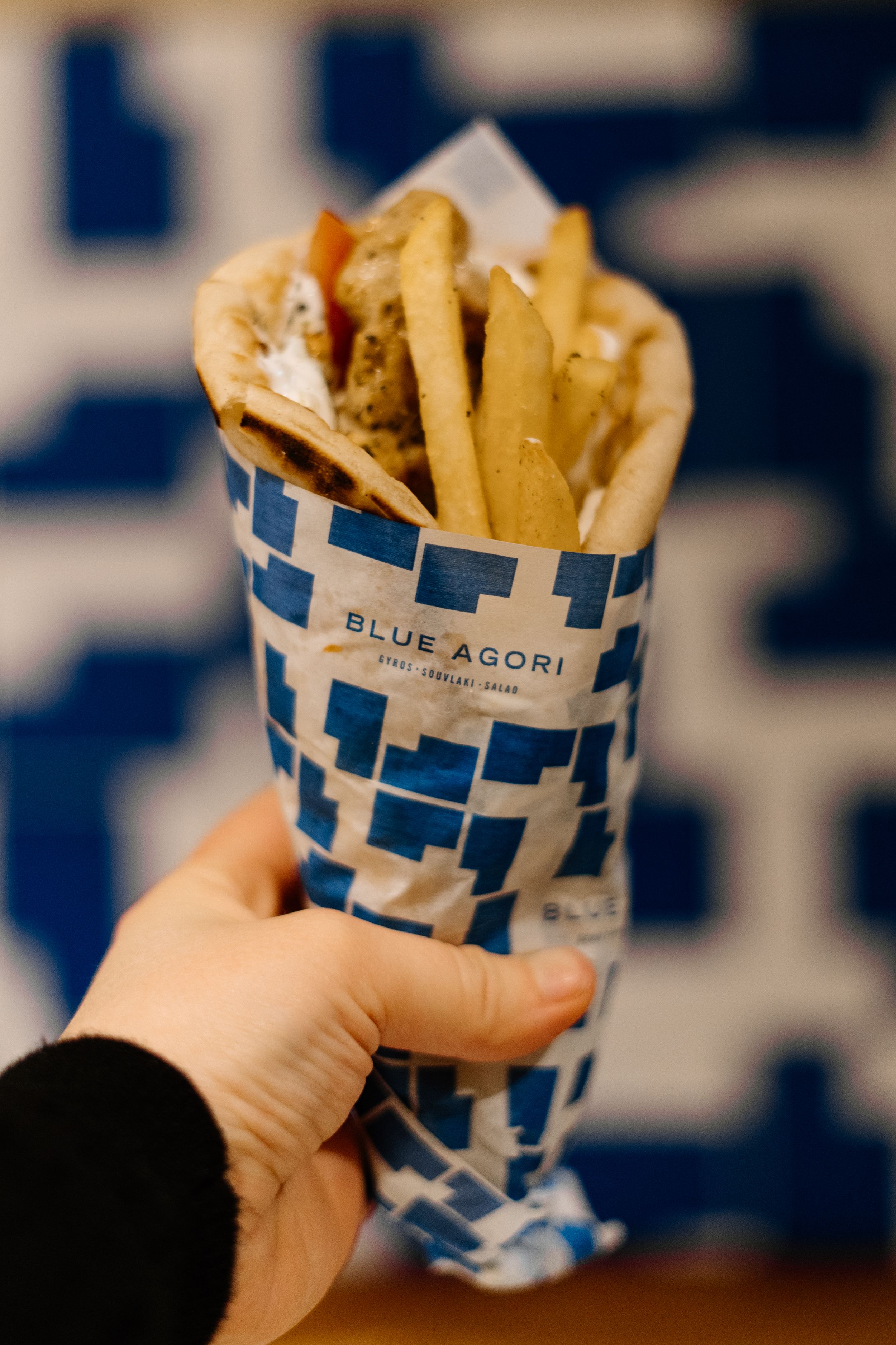 Hand holding a blue and white patterned paper cone filled with golden french fries from Blue Agori restaurant.