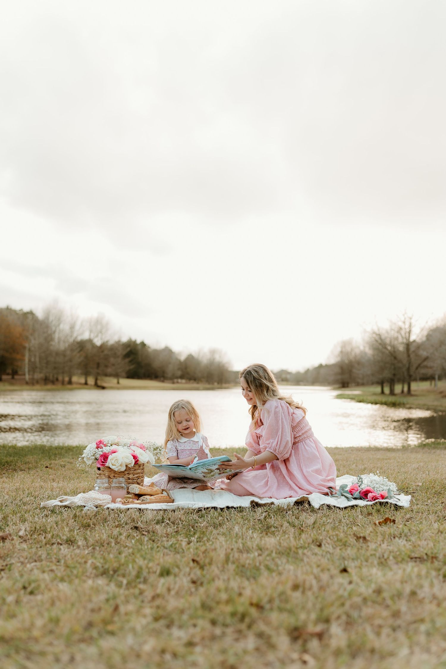 Peaceful lakeside picnic scene with pink flowers and two people relaxing on a blanket.