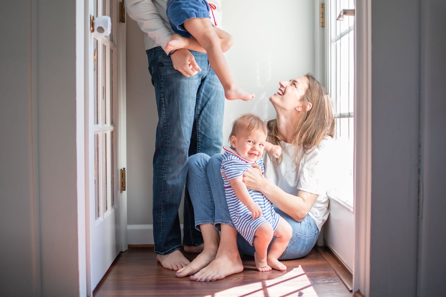 Family playfully sits together in a bright hallway with sunlight streaming through window.