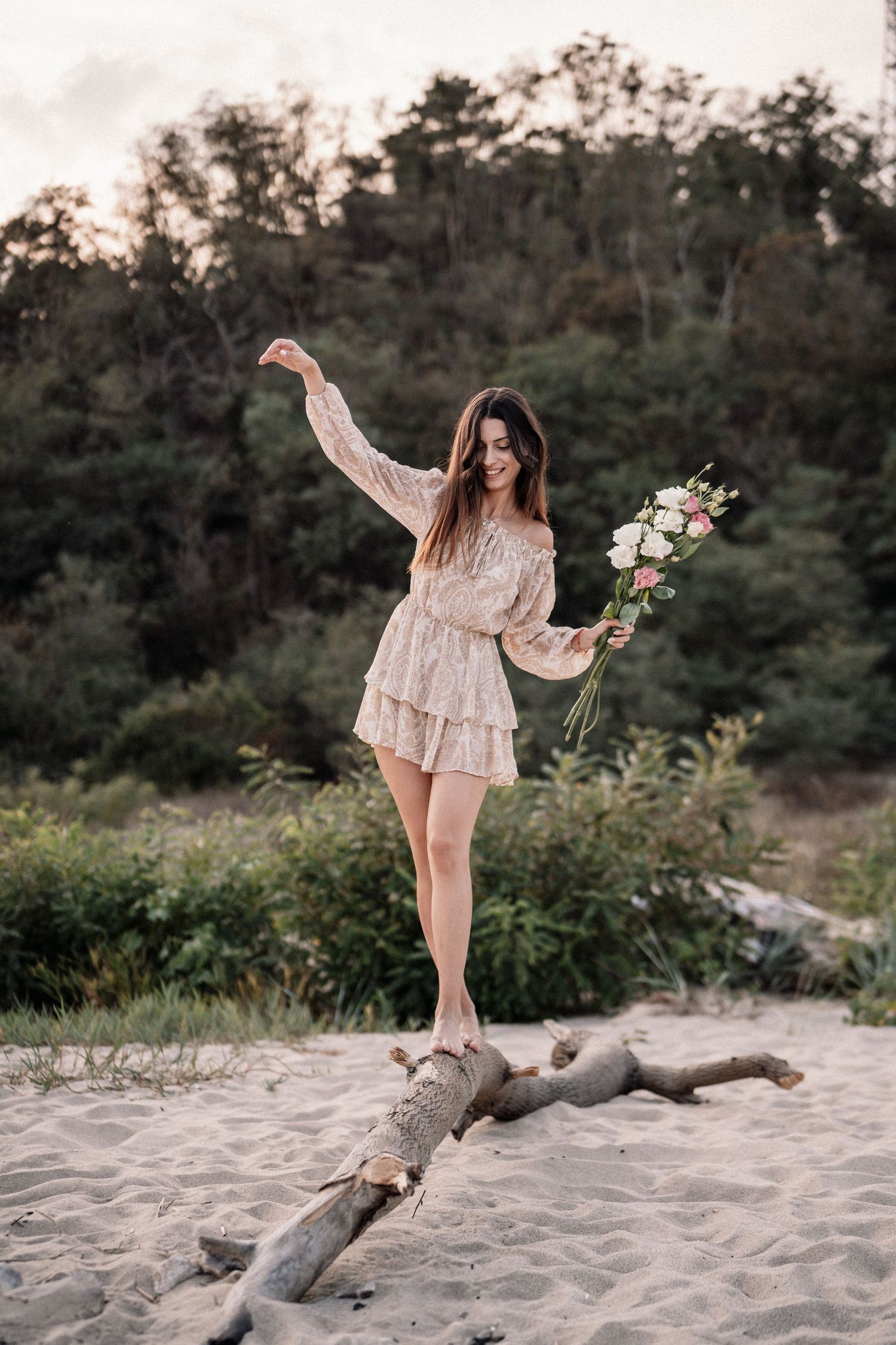 Graceful figure in ruffled dress balancing on driftwood with flowers on sandy beach.