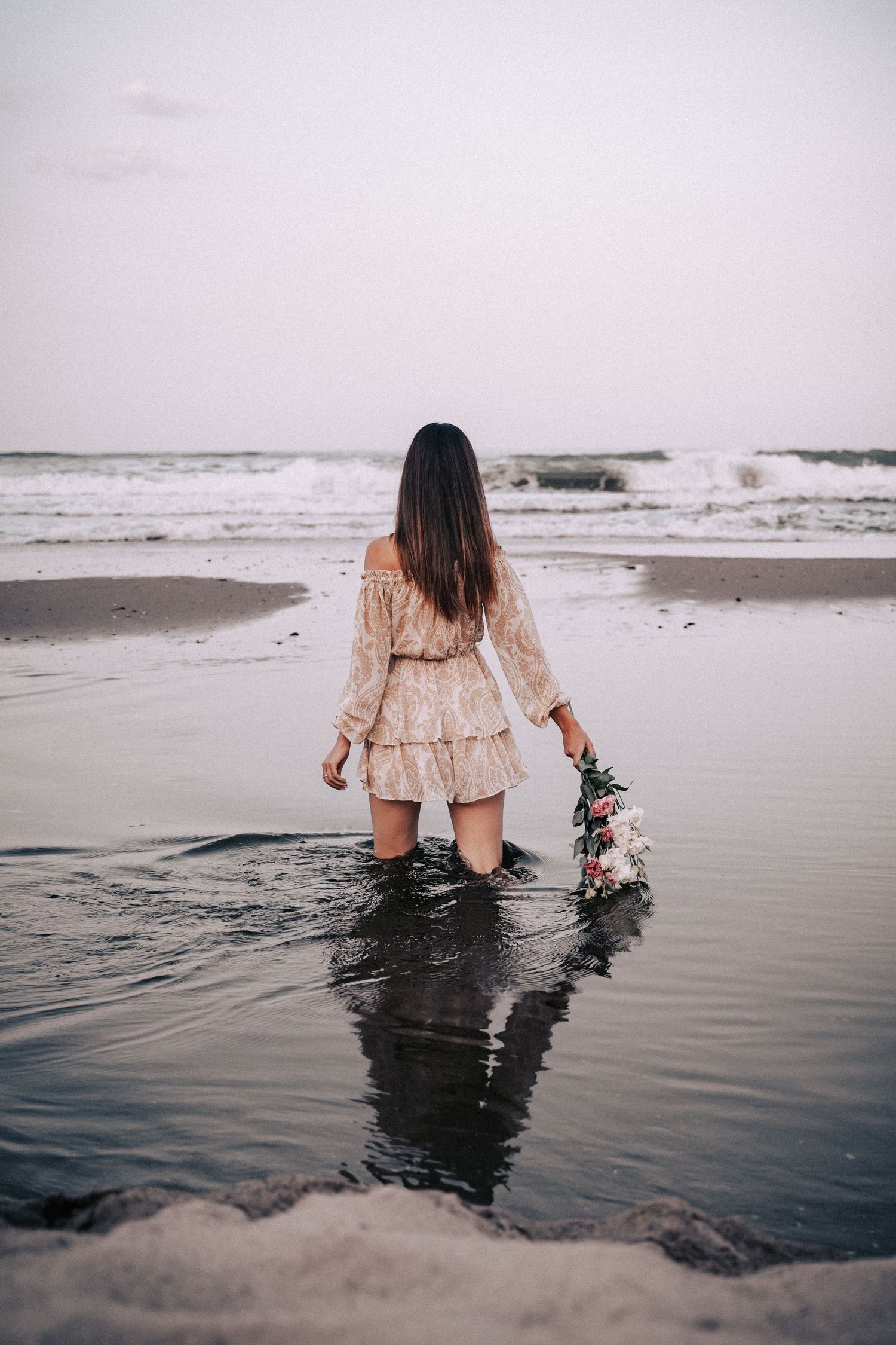 Person in flowing dress standing in shallow ocean waves with floral bouquet.