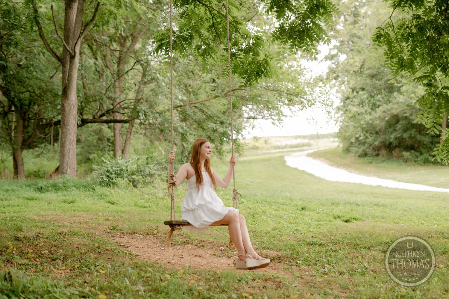 Graduate in white dress sits on wooden swing in peaceful wooded setting.