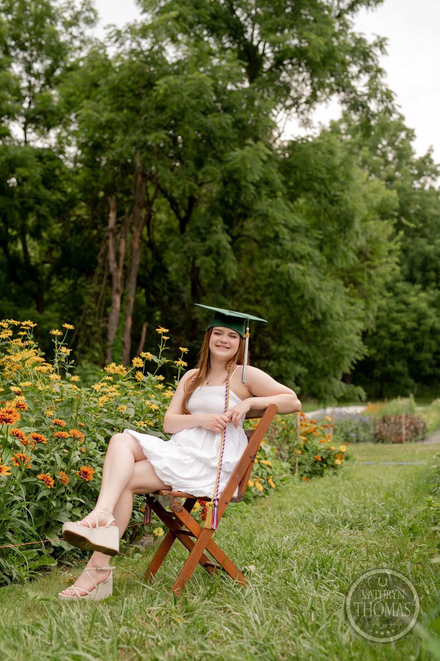 Graduate in white dress and cap relaxes in wooden chair near orange marigolds and forest backdrop.