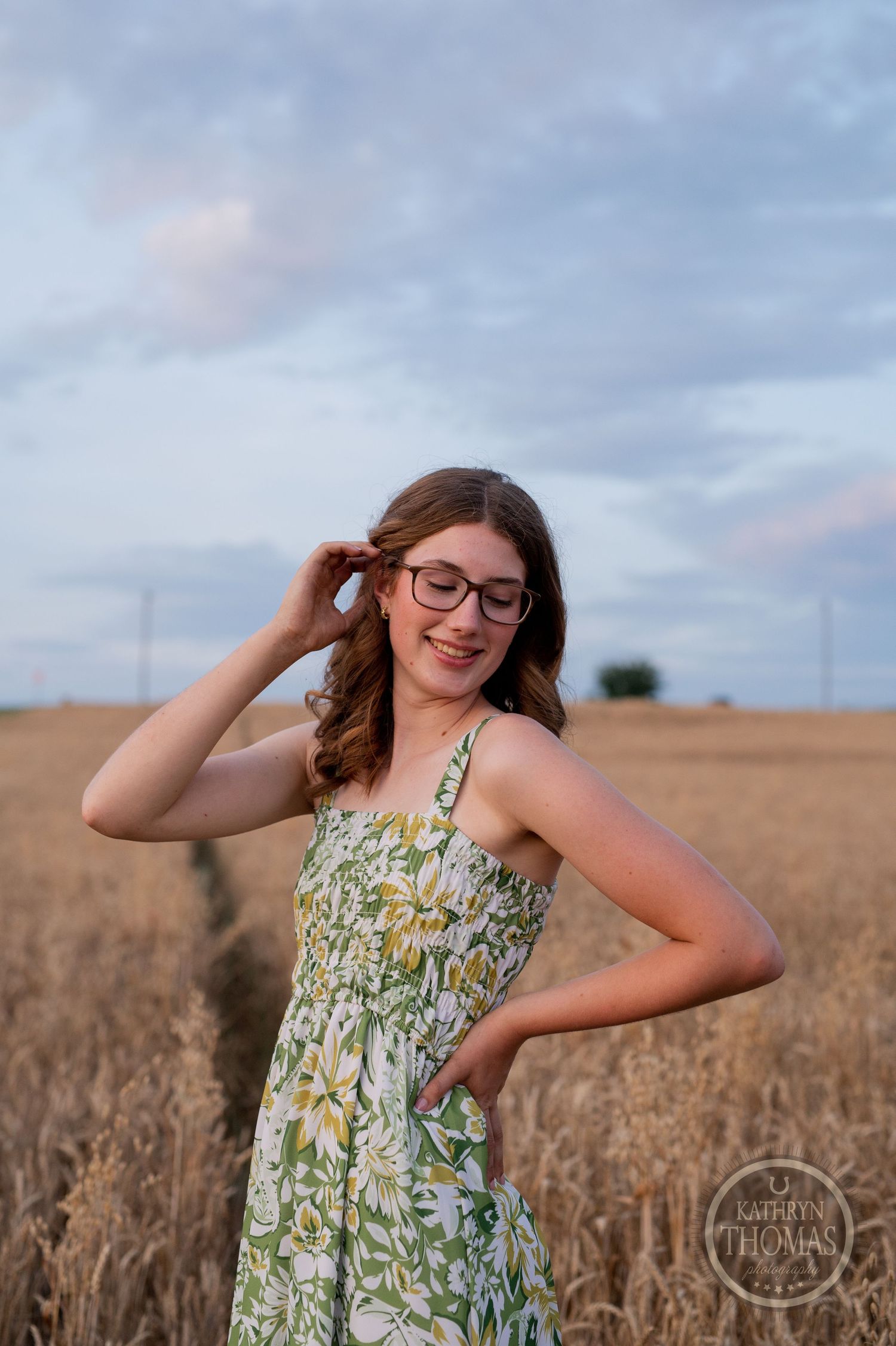 Person in floral dress and glasses poses playfully in wheat field during sunset.