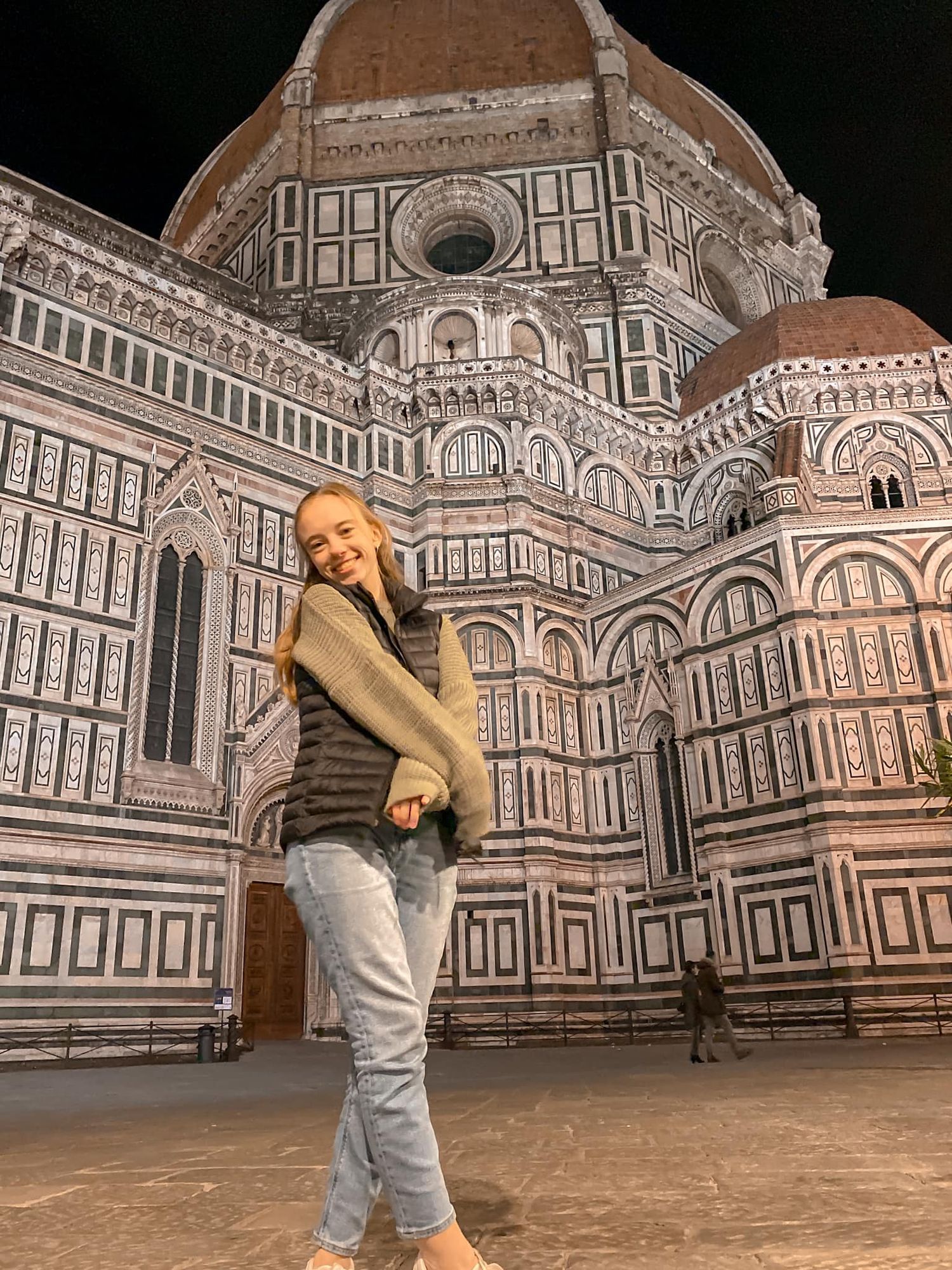 A tourist poses in front of the ornate facade of Florence's Duomo cathedral at night with its iconic Renaissance architecture.