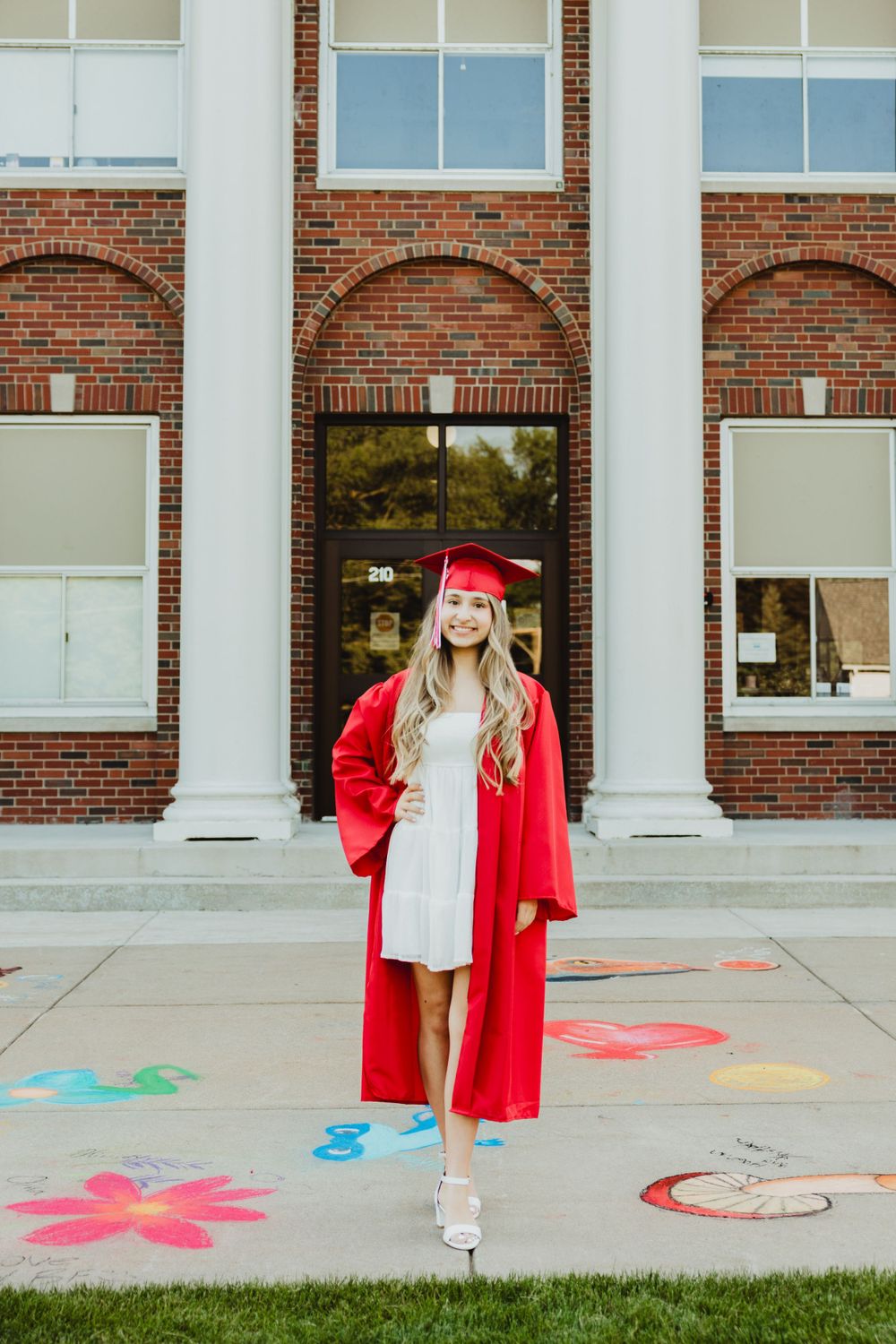 Graduate in red regalia poses near colorful chalk drawings on sidewalk outside brick building.