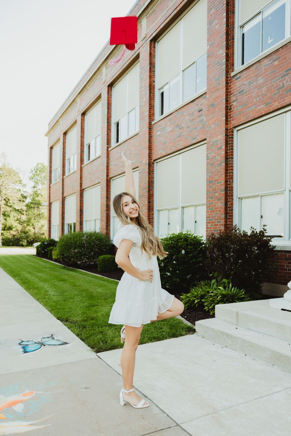 Playful graduate in red gown jumps and dances on sidewalk near brick building.