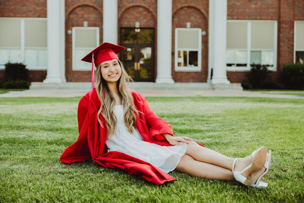 Graduate in red cap and gown poses on grassy lawn in front of brick building during graduation photoshoot.