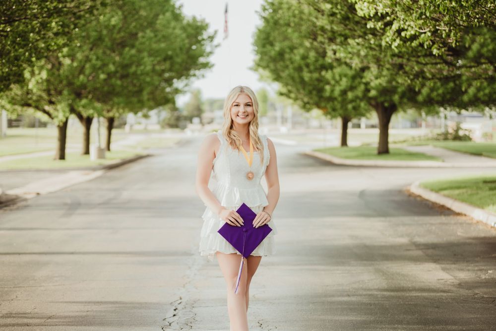 Graduate in white dress poses on tree-lined street during sunny outdoor photo session.