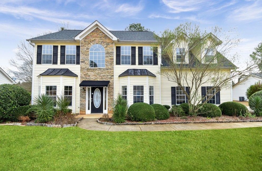 Two-story traditional suburban home with white siding, black shutters and stone accents on a manicured green lawn.