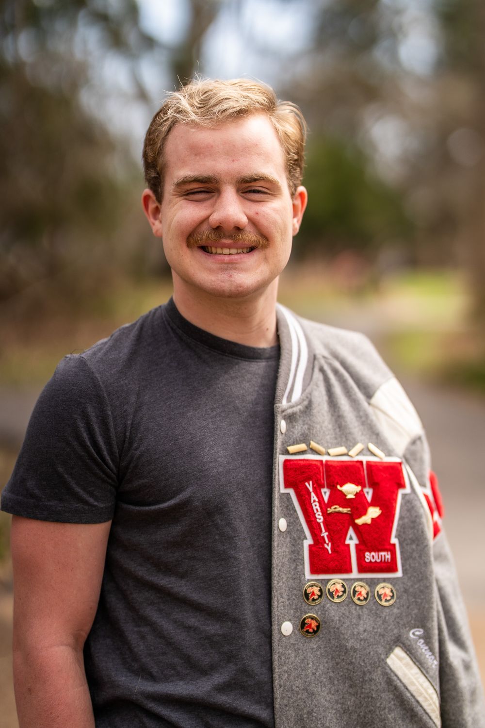 Young man smiling outdoors wearing a gray t-shirt and letterman jacket with red W patch and pins.