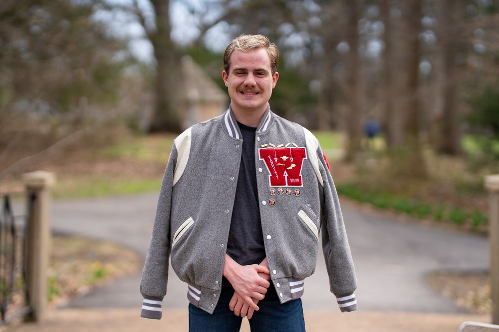 Young man smiling outdoors wearing a gray varsity jacket with red letter, standing on a tree-lined path.