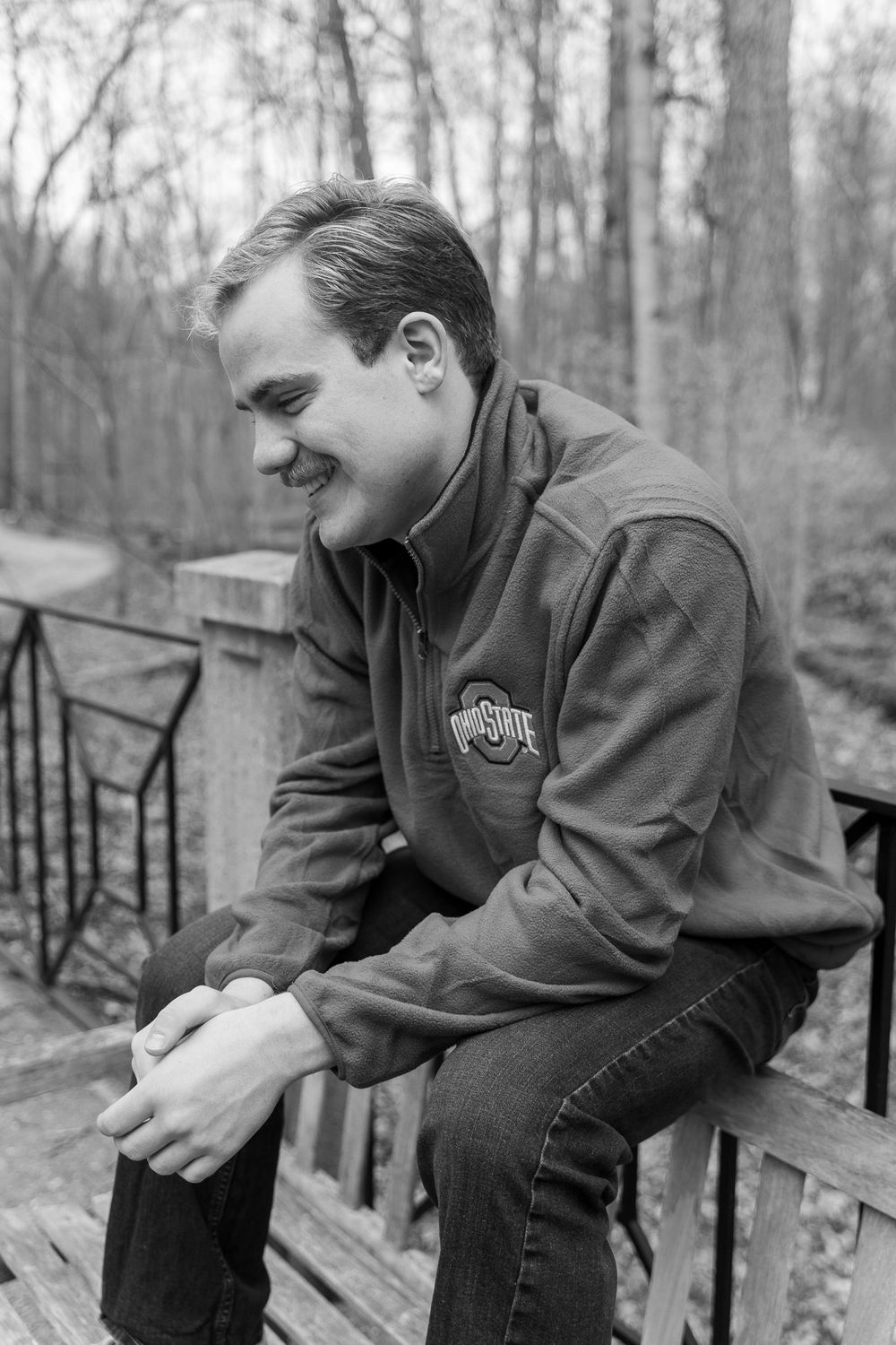 Young man in Ohio State fleece jacket smiling while sitting on wooden bridge in black and white photo.