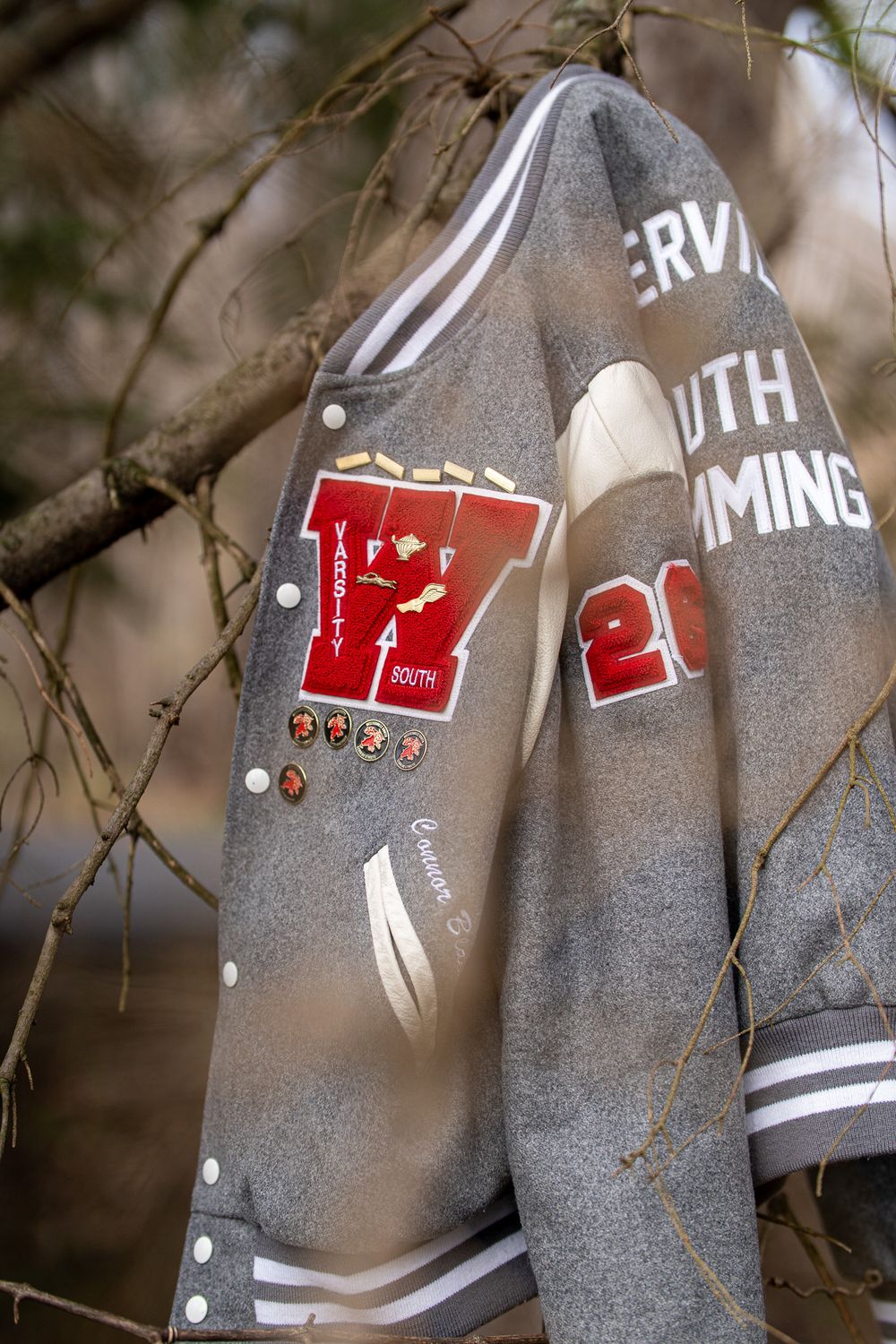 Gray varsity letterman jacket with red W logo and swim team patches hanging on tree branches outdoors.