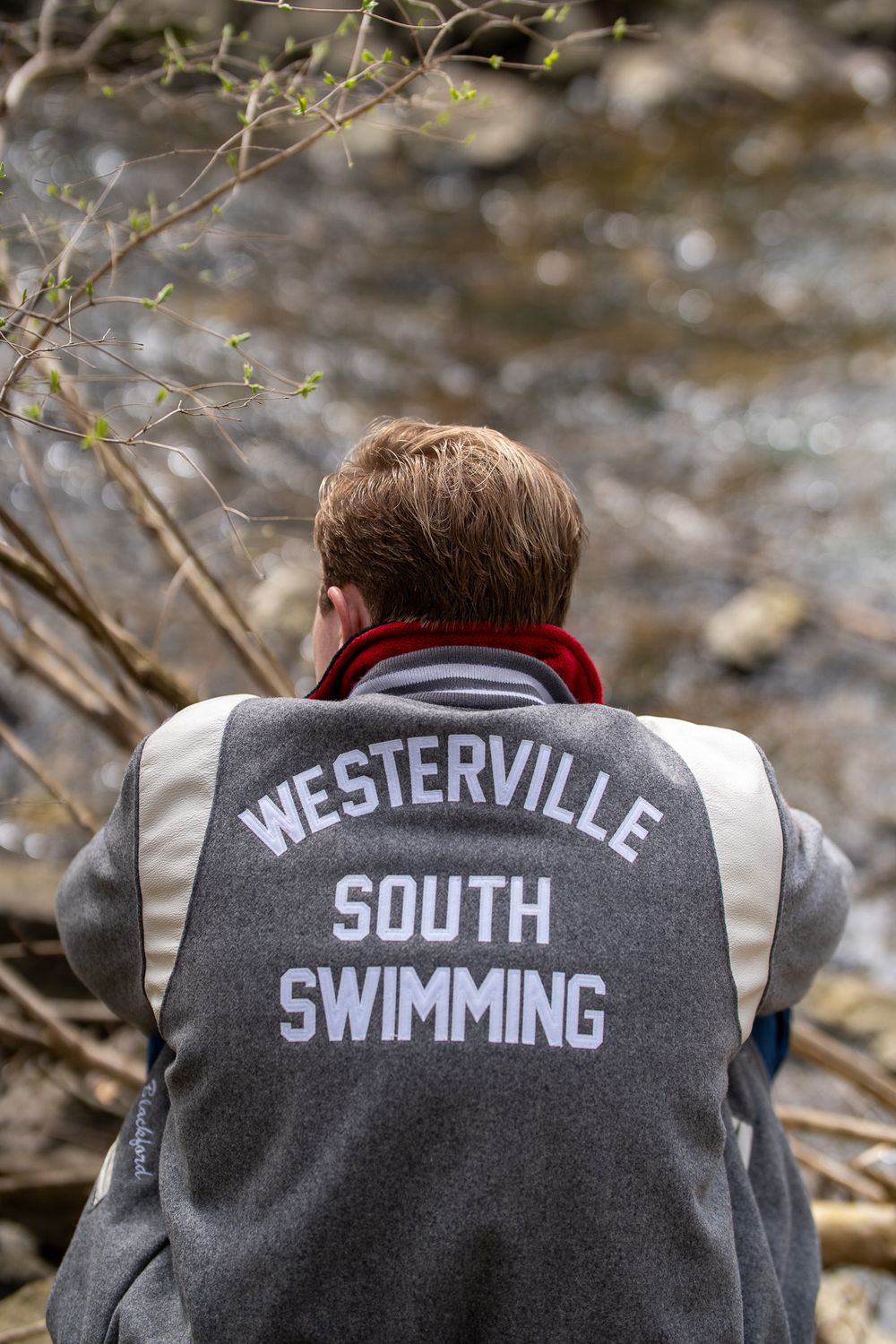 Person wearing a Westerville South Swimming jacket standing near a rocky stream outdoors.