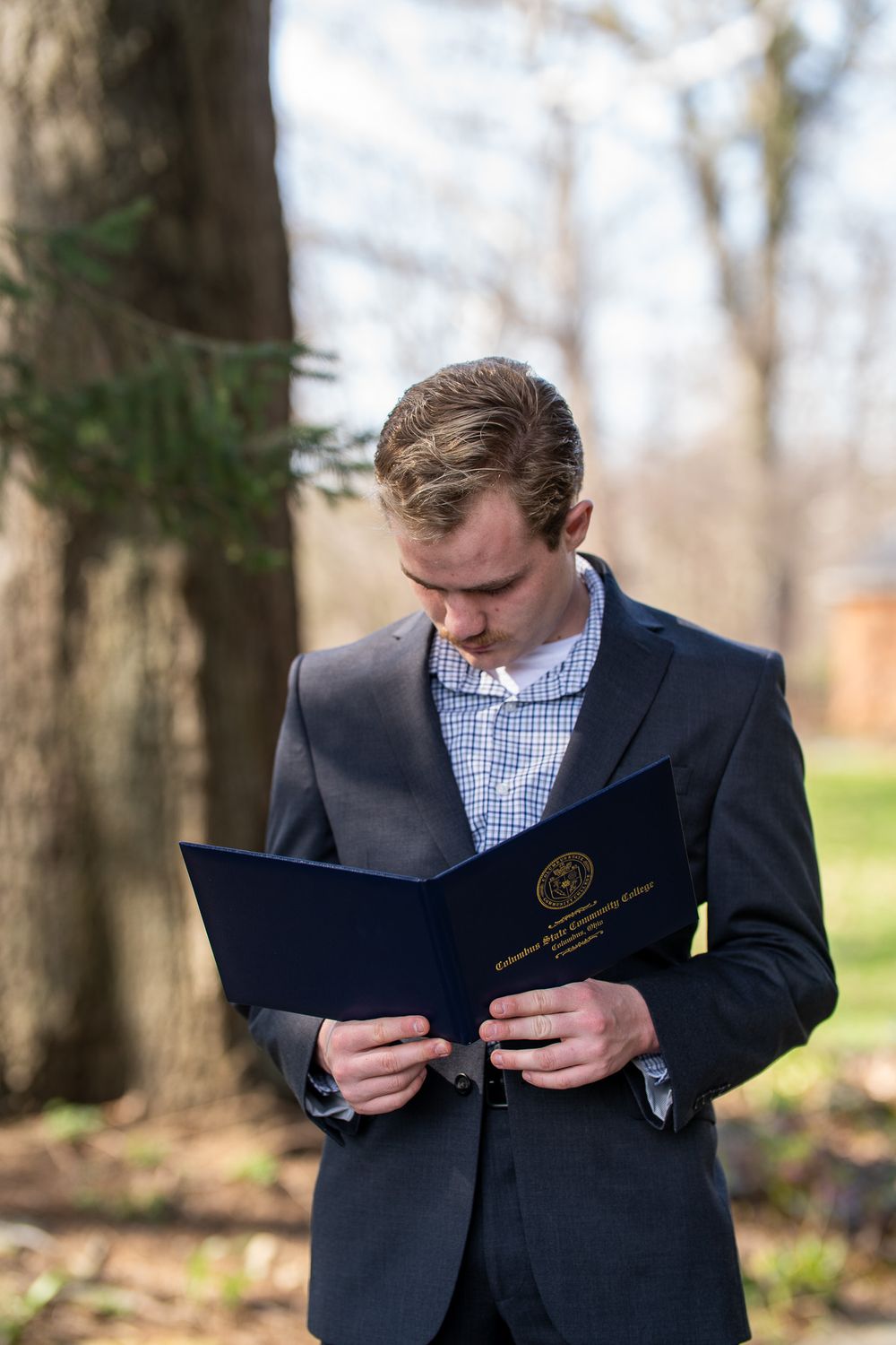 Young man in dark suit reading blue diploma folder outdoors among trees in natural setting.