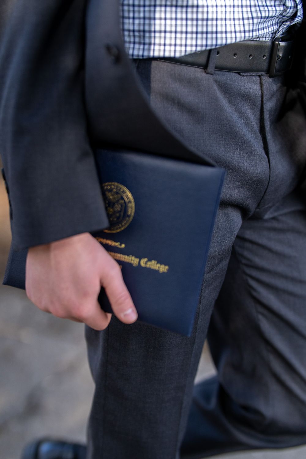 Man in suit holding a navy blue Community College diploma cover at graduation ceremony.