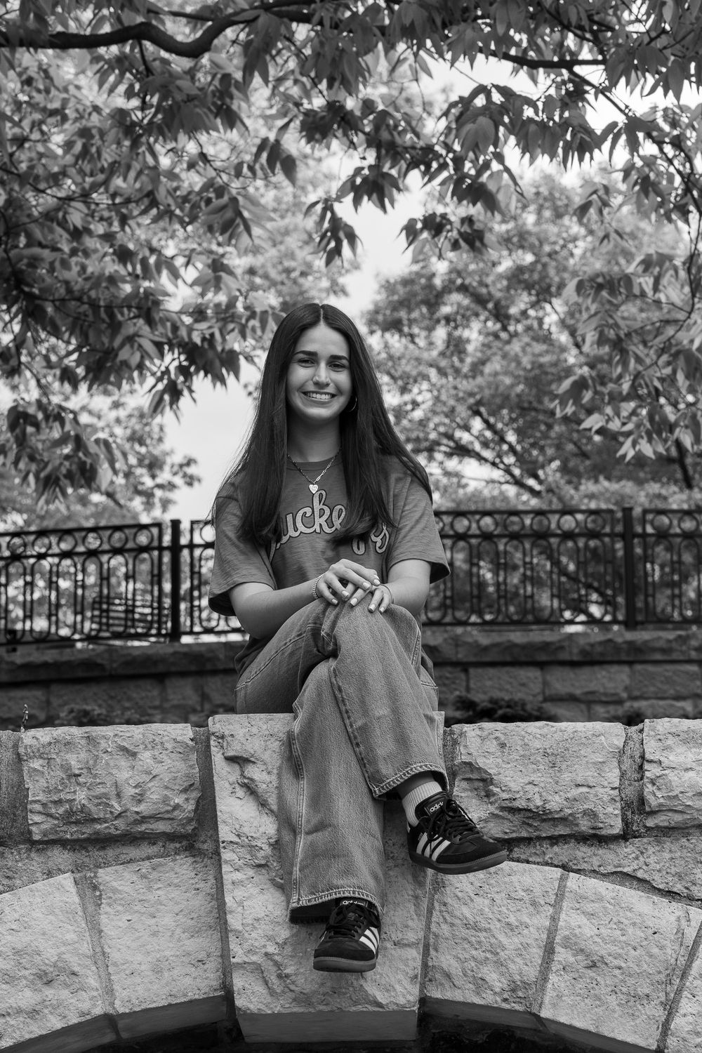 Young woman smiling, seated on stone wall outdoors, wearing jeans and t-shirt, surrounded by trees, black and white photo.