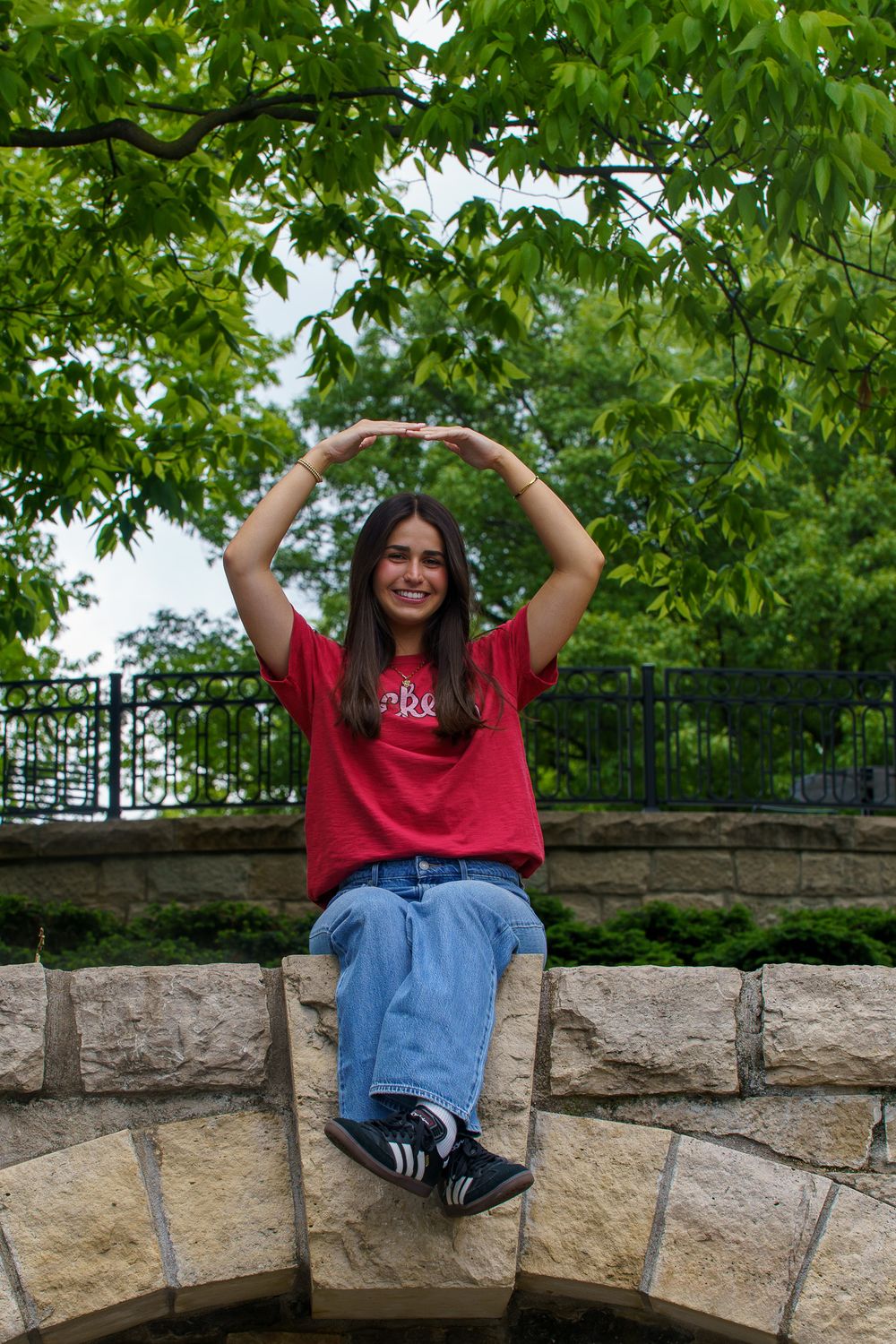 Young woman in red t-shirt and jeans sits on stone wall, arms raised overhead, surrounded by lush green trees.