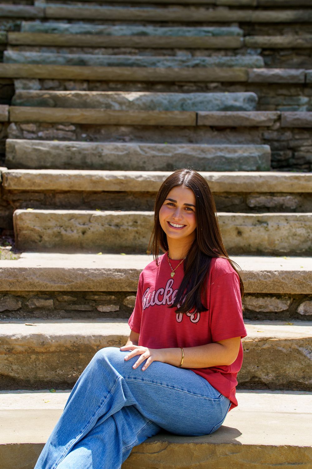 Young woman in red Buckeyes tee and jeans smiling while sitting on stone steps outdoors in sunlight.