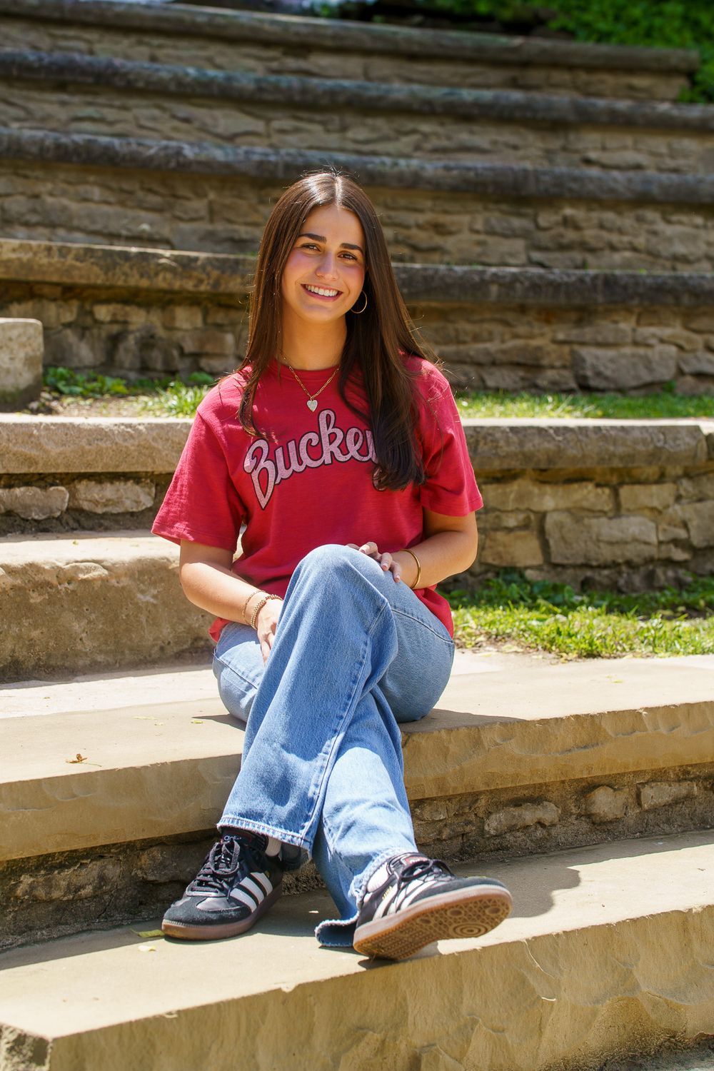 Smiling young woman in red Buckeyes shirt and jeans sitting on stone steps outdoors.