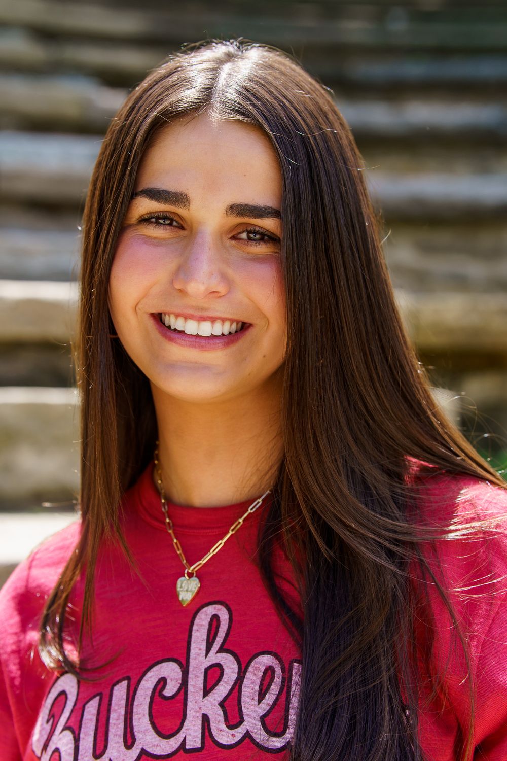 Smiling young woman with long dark hair wearing a red Buckeyes shirt and gold heart necklace outdoors.