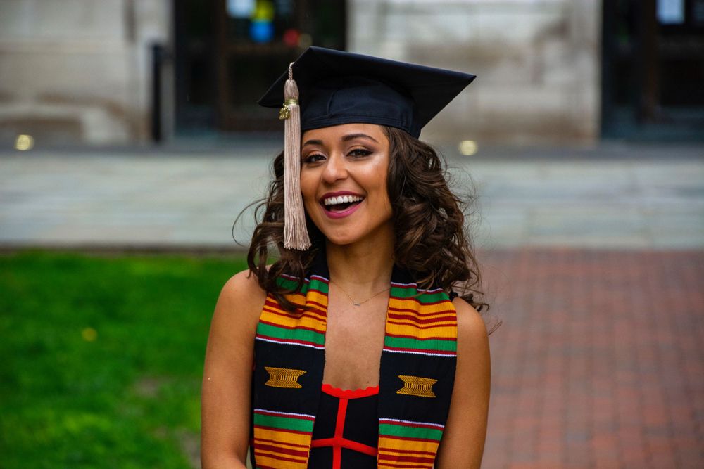 Graduate in cap and gown with colorful stole posing outdoors on campus.