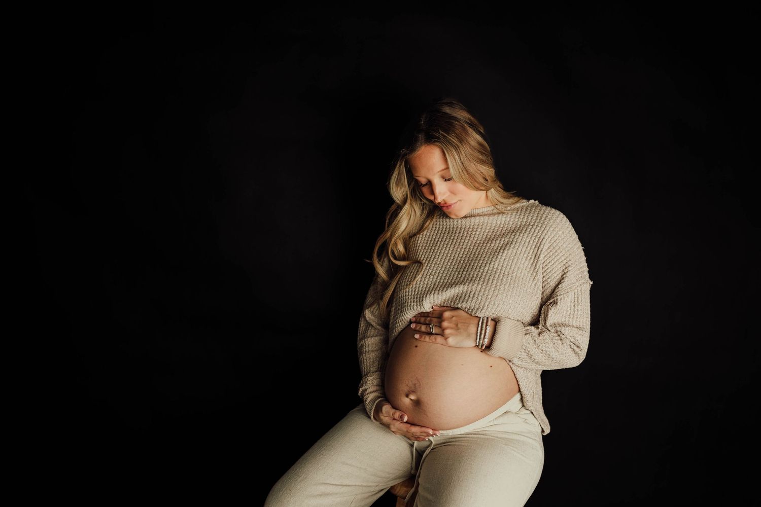 Maternity photo of pregnant woman in beige sweater against black background.