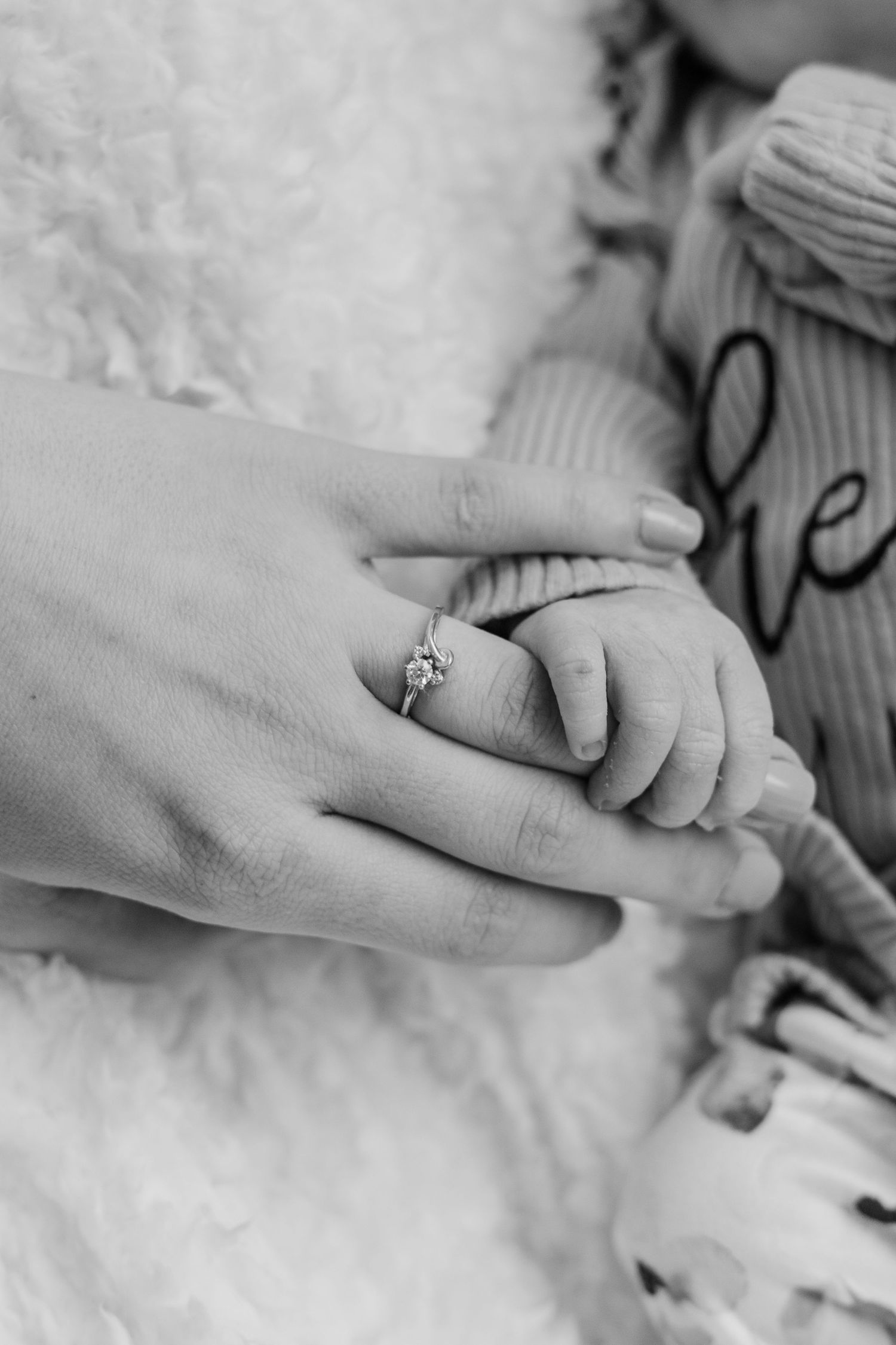 Close-up black and white photo of an adult's hand holding a baby's hand with an engagement ring visible.