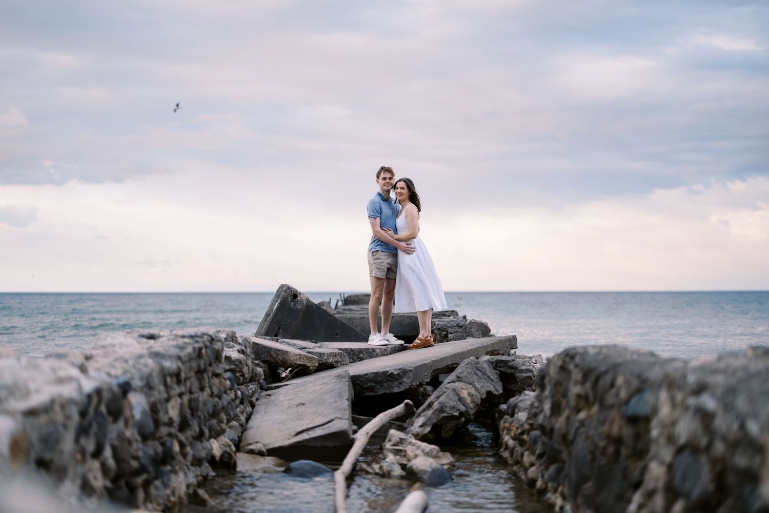 A romantic couple stands together on coastal rocks at the beach during sunset with ocean stretching to the horizon.