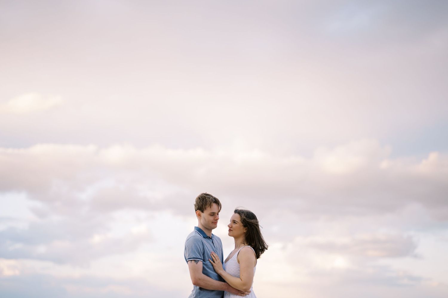 Romantic couple embraces against a dreamy pink sunset sky with wispy clouds in the background.