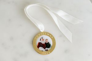 A round gold ornament with a photo insert hangs from a white satin ribbon against a light background.