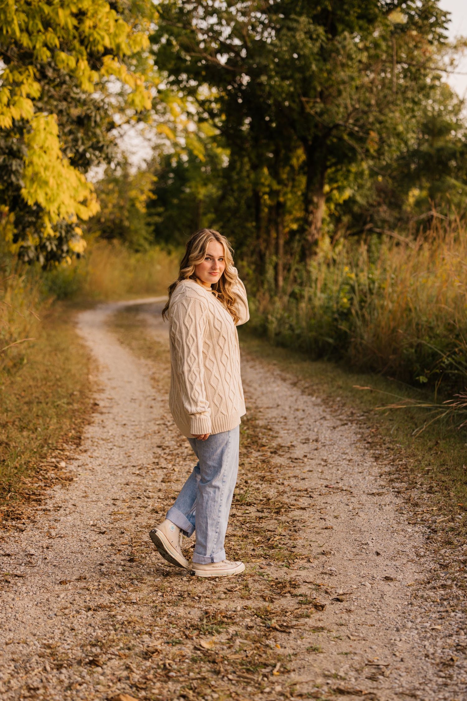Person in cozy cream cable knit sweater walking along serene autumn nature trail at golden hour.
