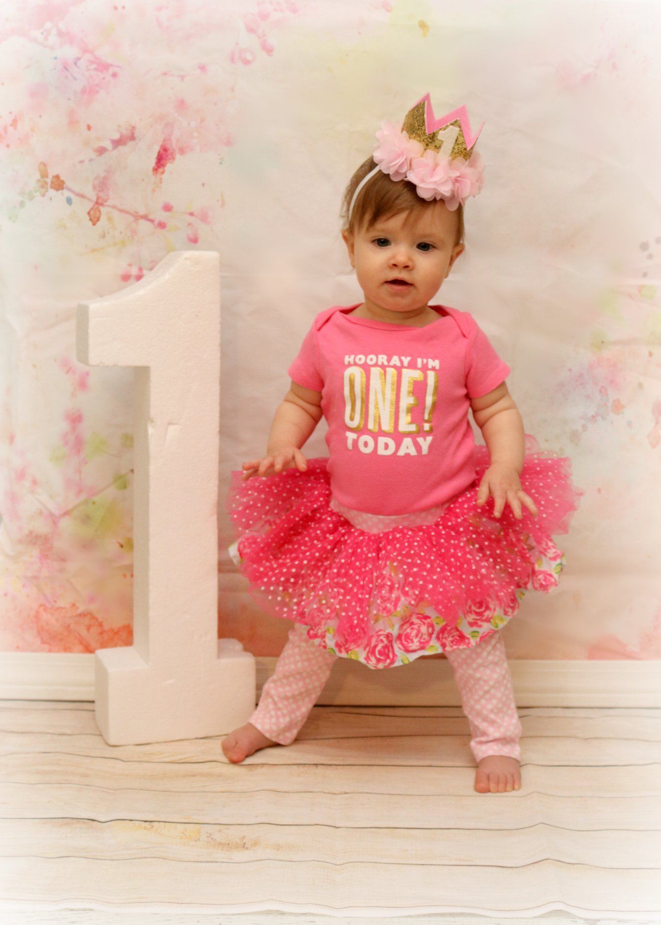 Baby wearing pink tutu and crown poses with large number one prop.