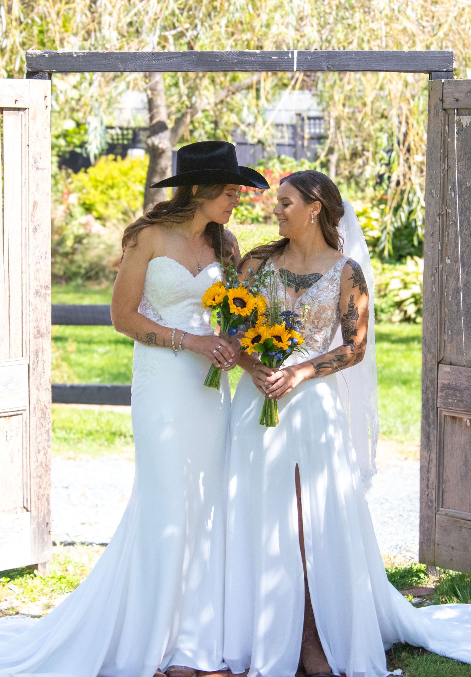 Two brides in white gowns share a moment under a wooden arch in a garden setting.