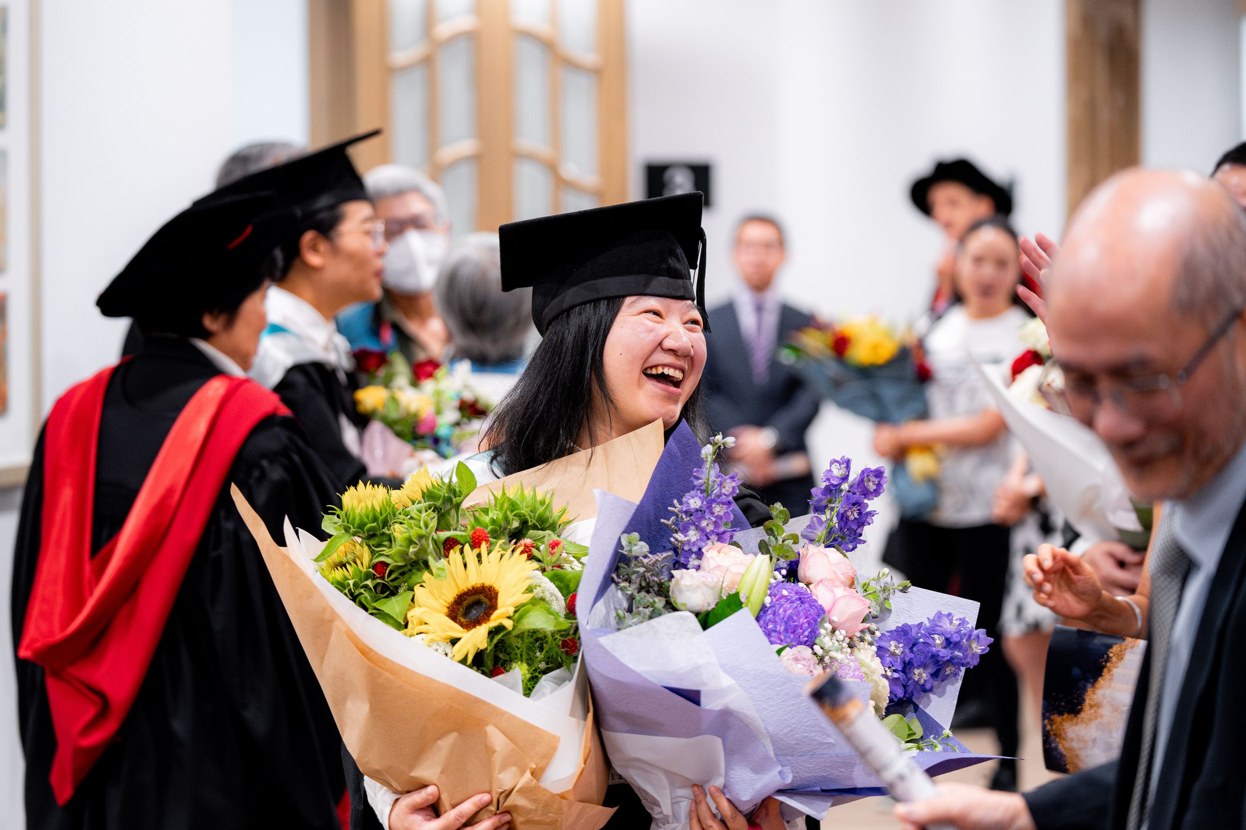 Graduate holding sunflower bouquet laughs during post-ceremony celebrations.