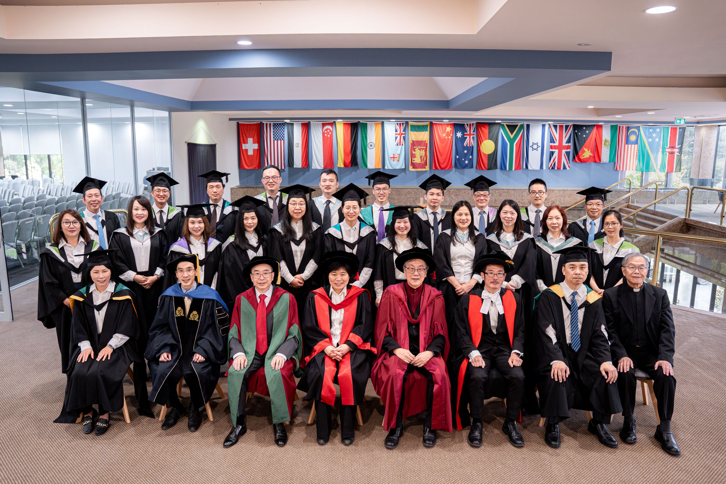 Large group of graduates in academic regalia seated together with international flags displayed on the wall.