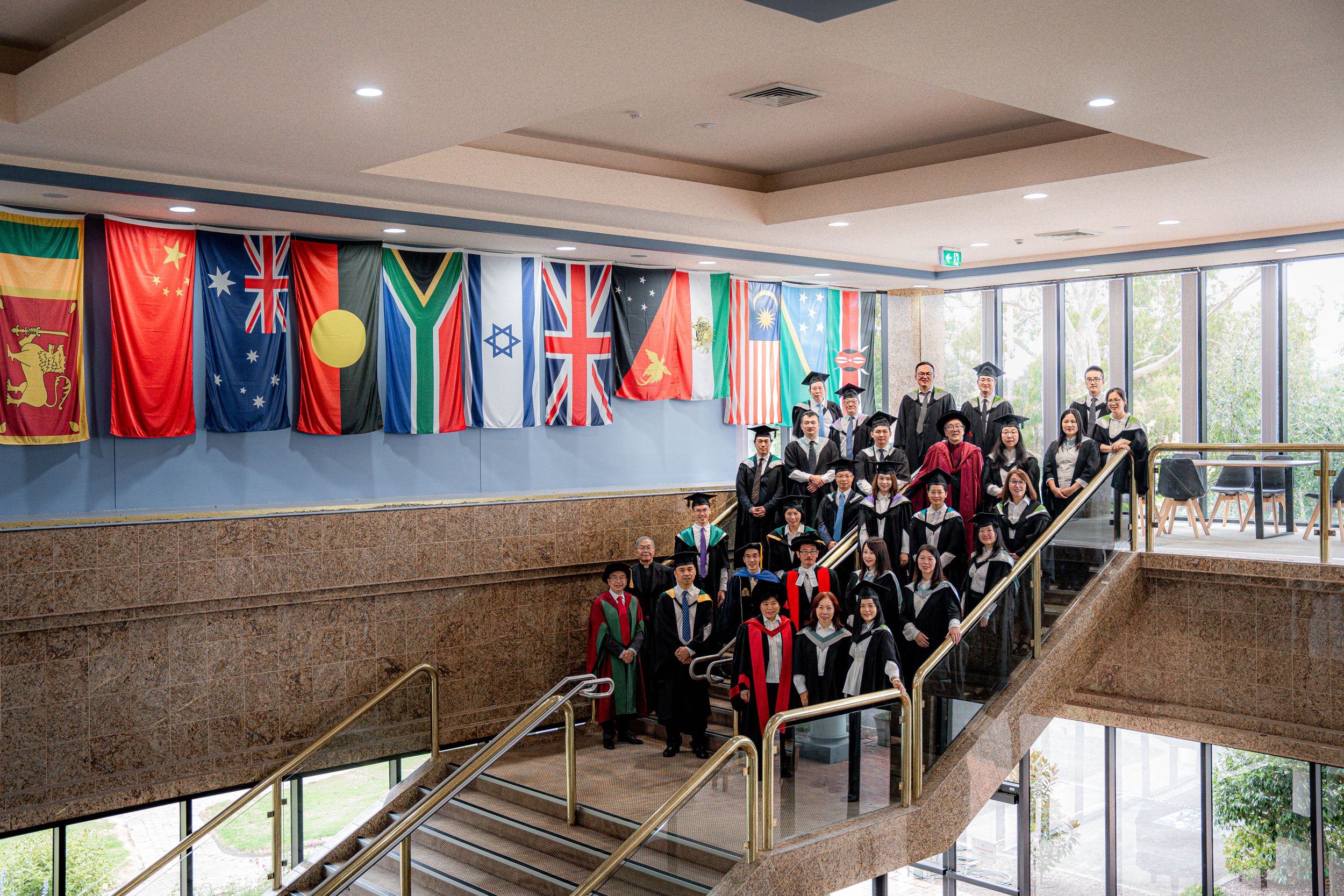 Graduates in academic dress gathered on stairs with international flags displayed on the wall.