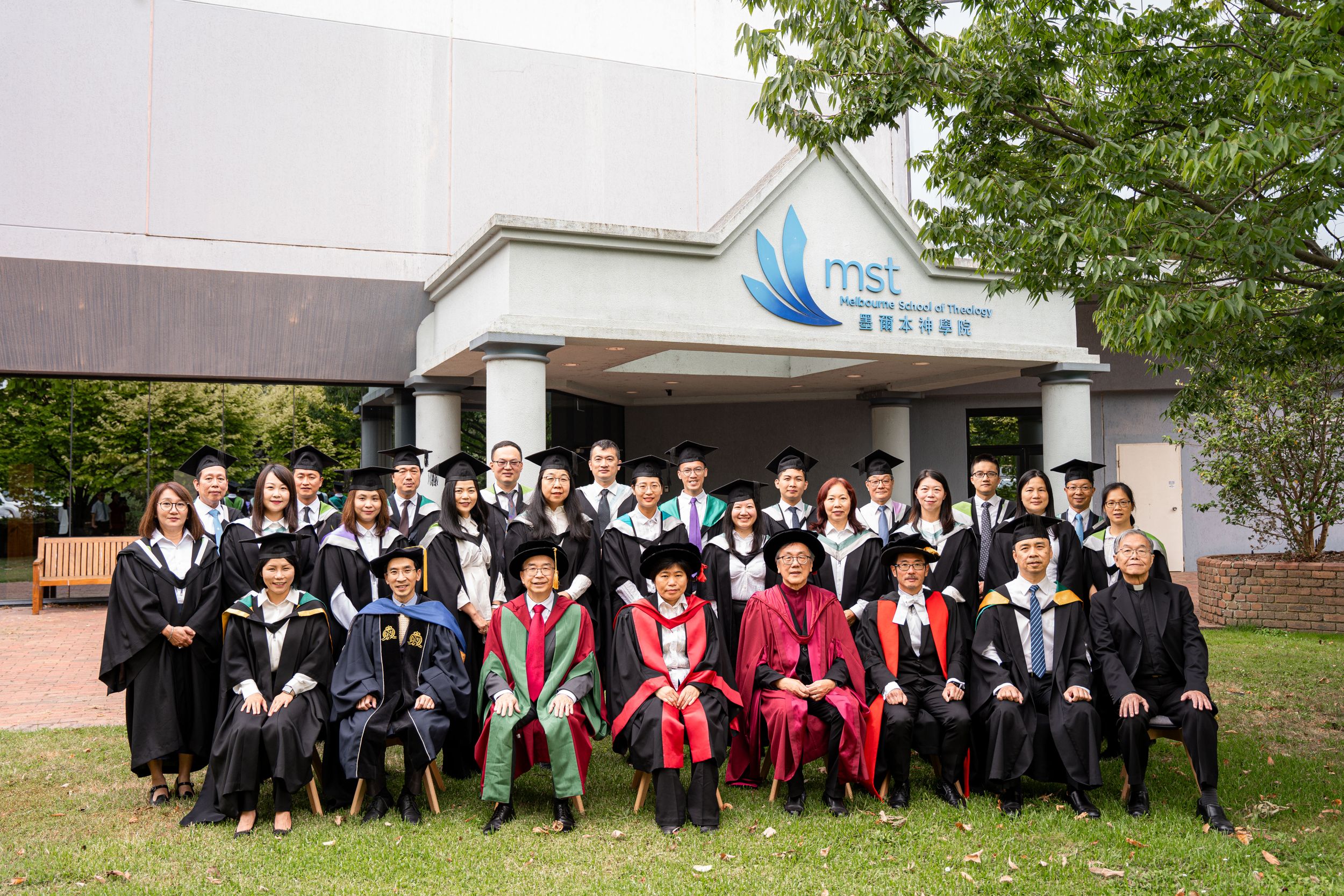 Graduates in academic regalia pose for group photo outside an academic building.