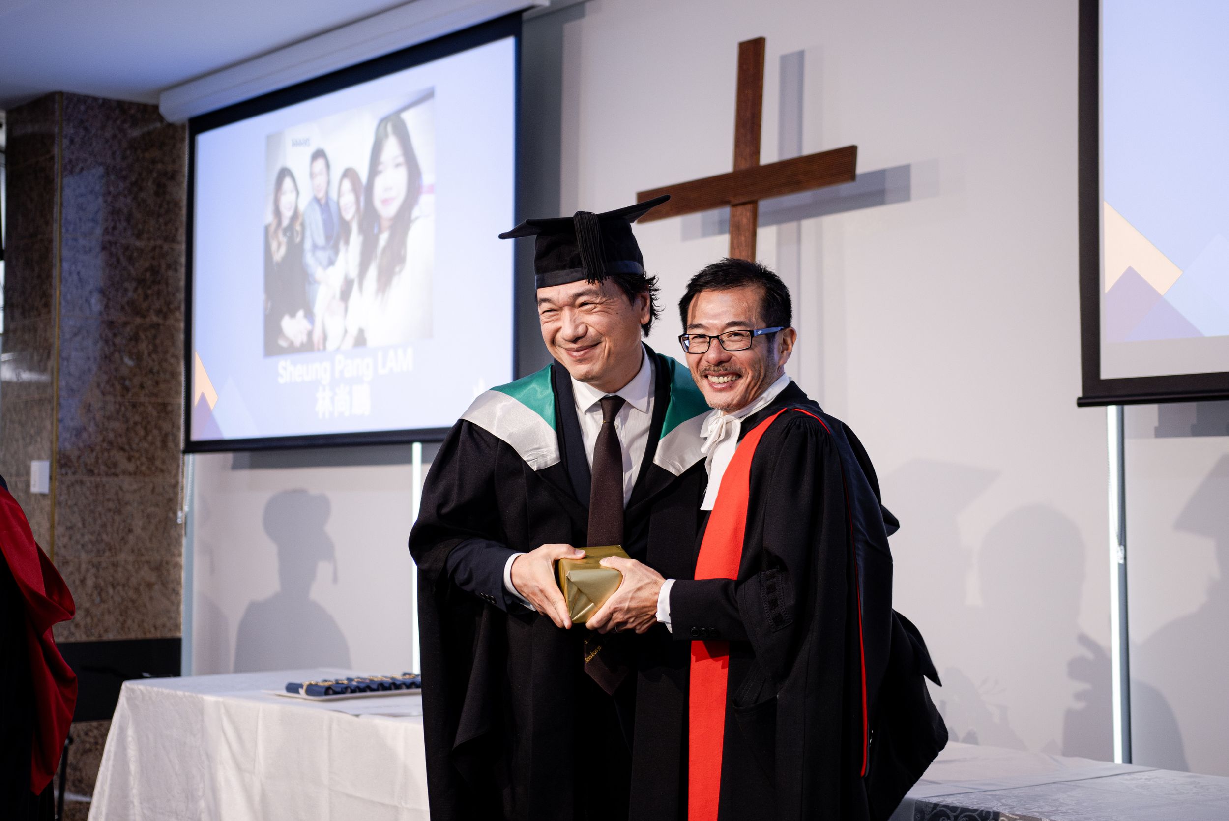 Academic ceremony with graduates in traditional regalia exchanging documents in front of a cross.