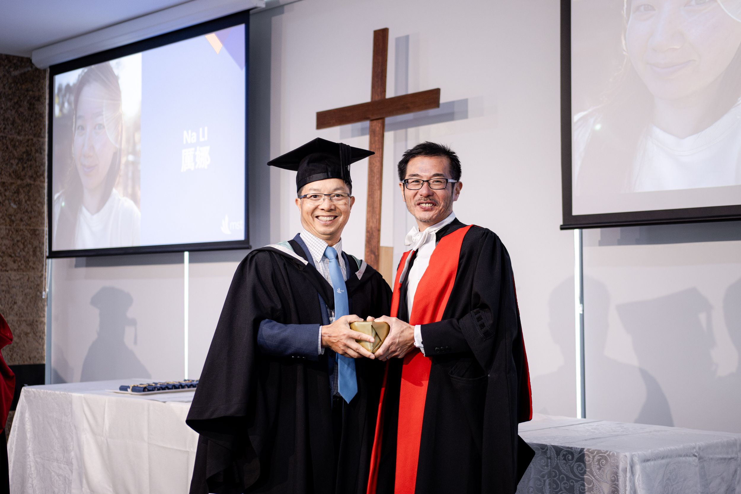 Graduation ceremony with diploma presentation in front of religious symbols and projector screens.