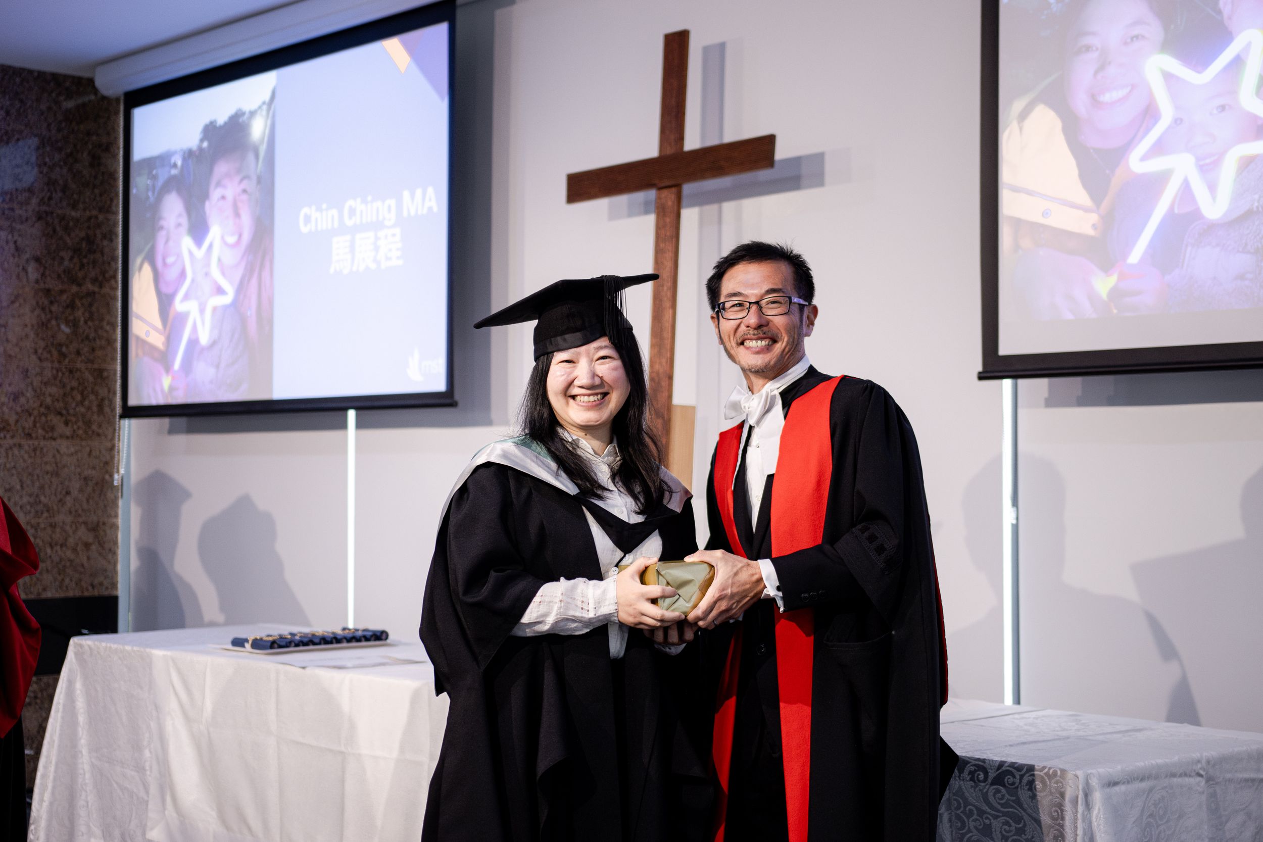 Graduate receiving diploma during ceremony with cross and projection screens in background.