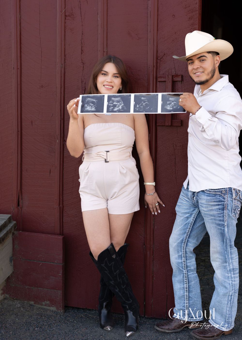 A couple poses together against a red wall while holding ultrasound photos.