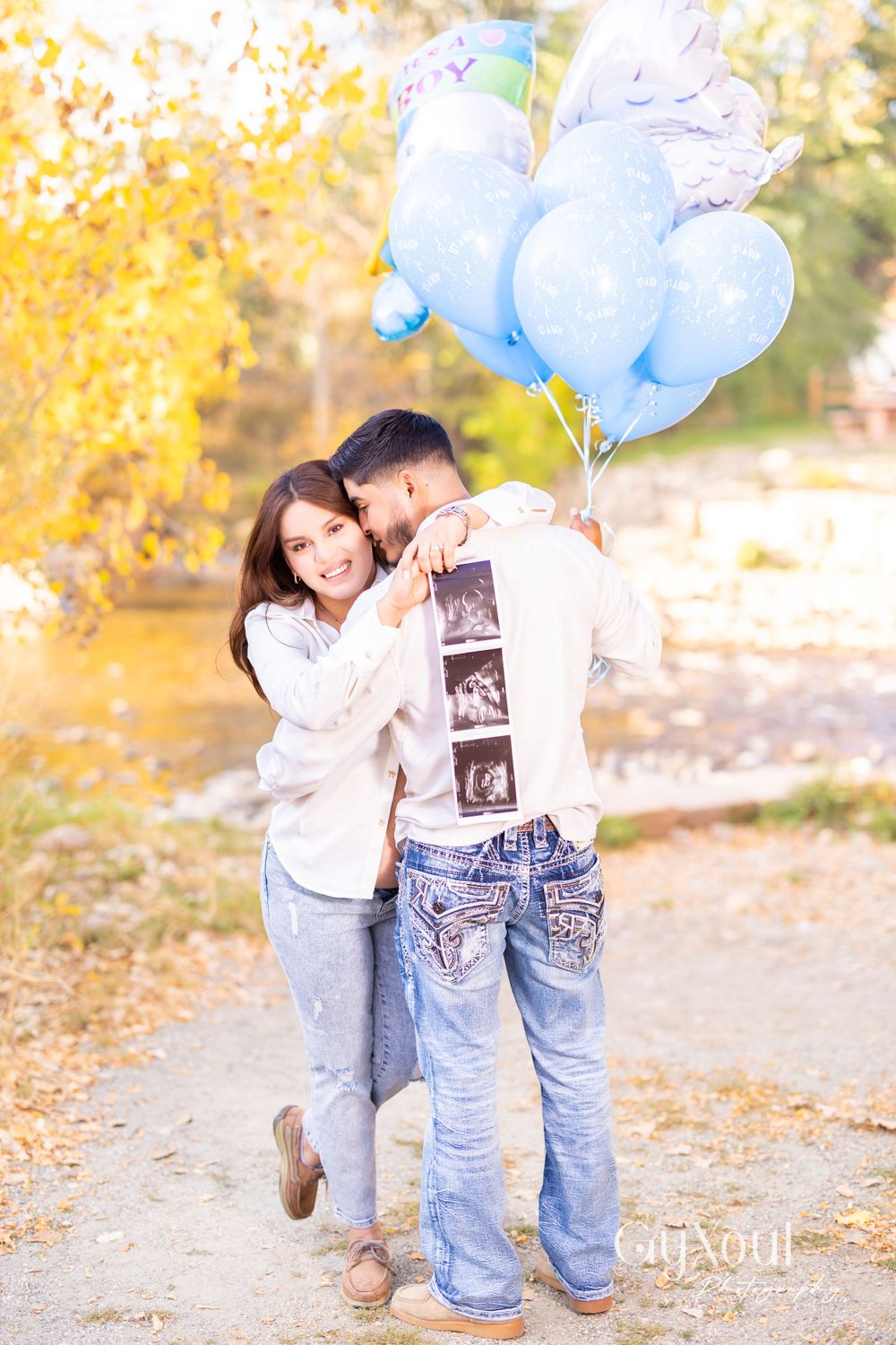 A couple embraces while holding ultrasound strips and blue balloons in an autumn setting.