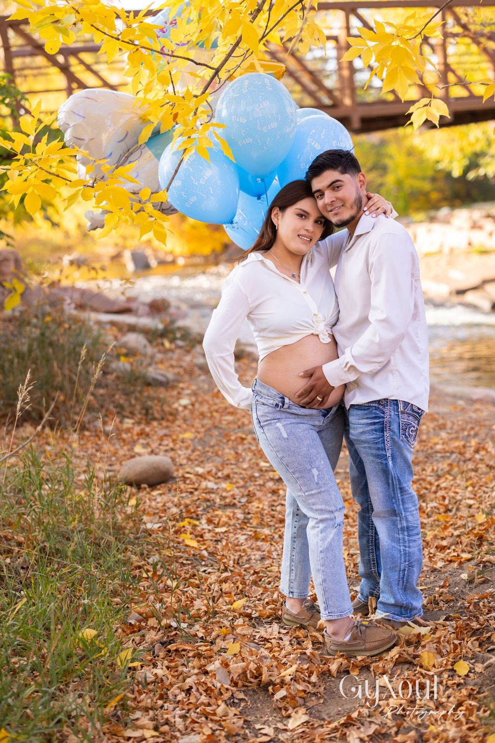 A maternity photo shoot in a fall setting with blue balloons and yellow leaves.