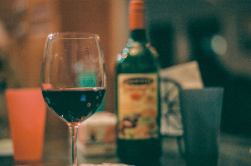 A glass of red wine sits on a table with a bottle in soft focus background lighting.